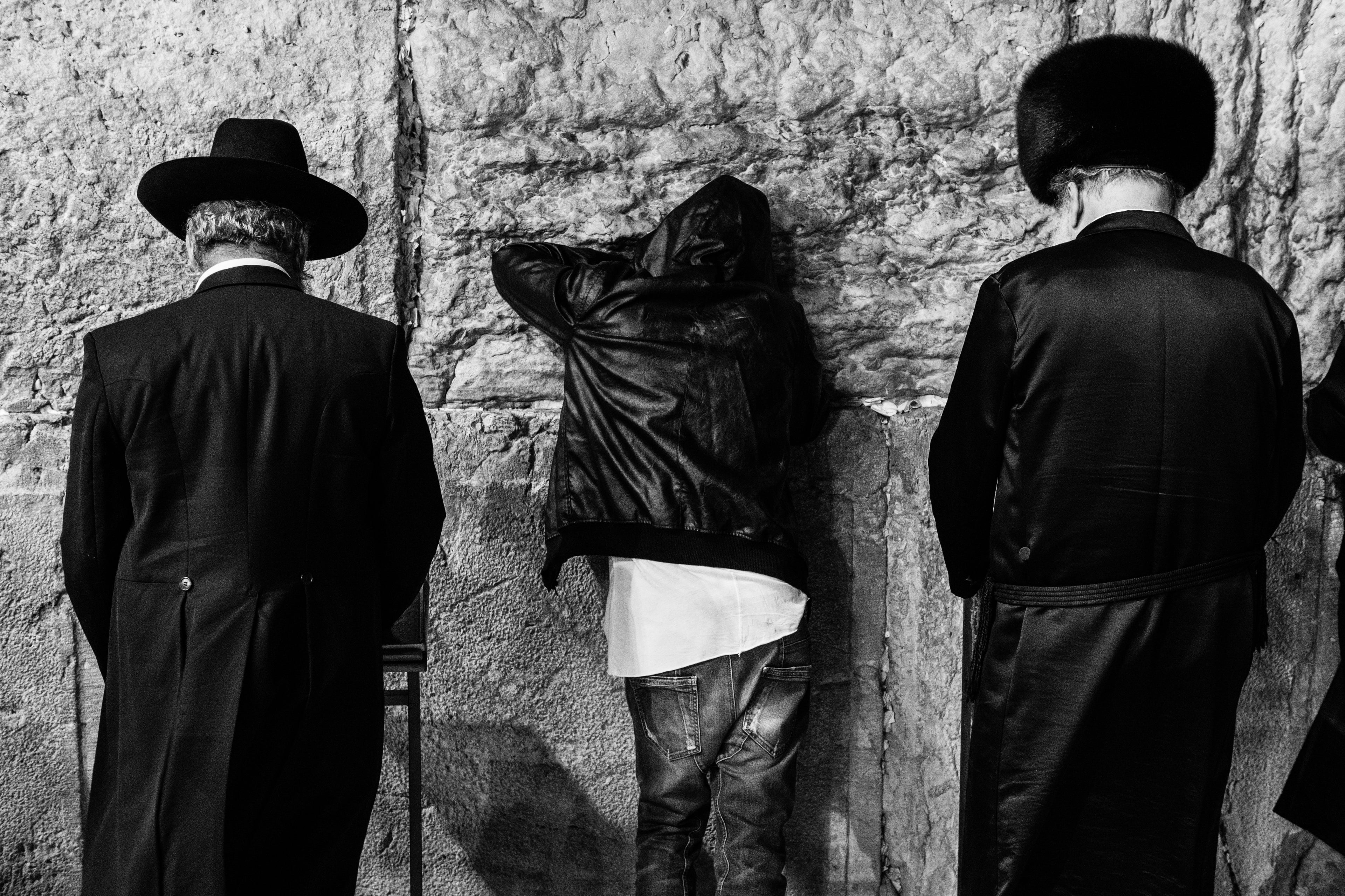 Black and white Kotel Western Wall of three men in traditional Jewish praying in front of a stone wall, with one man leaning against the wall in prayer.